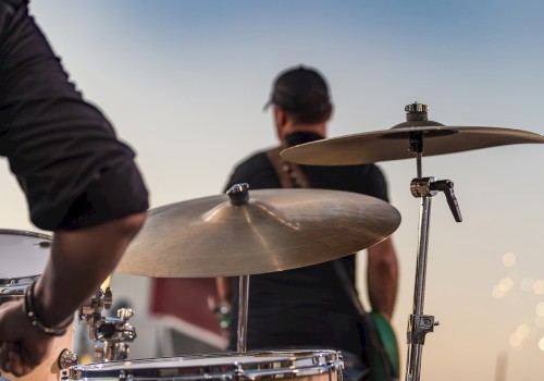 A drummer and a bassist perform outdoors at sunset, with the focus on the drum cymbals and blurred background.