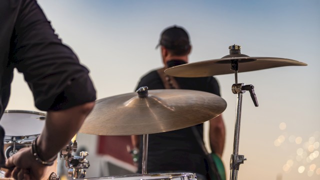 A drummer playing their instrument outdoors during sunset, with a person in the background and a calm, bokeh-lit sky.