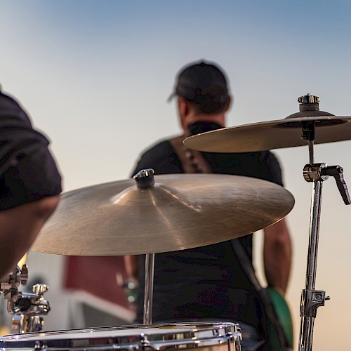 A person plays drums outdoors during sunset, with another musician in the background and bokeh lights in the distance.