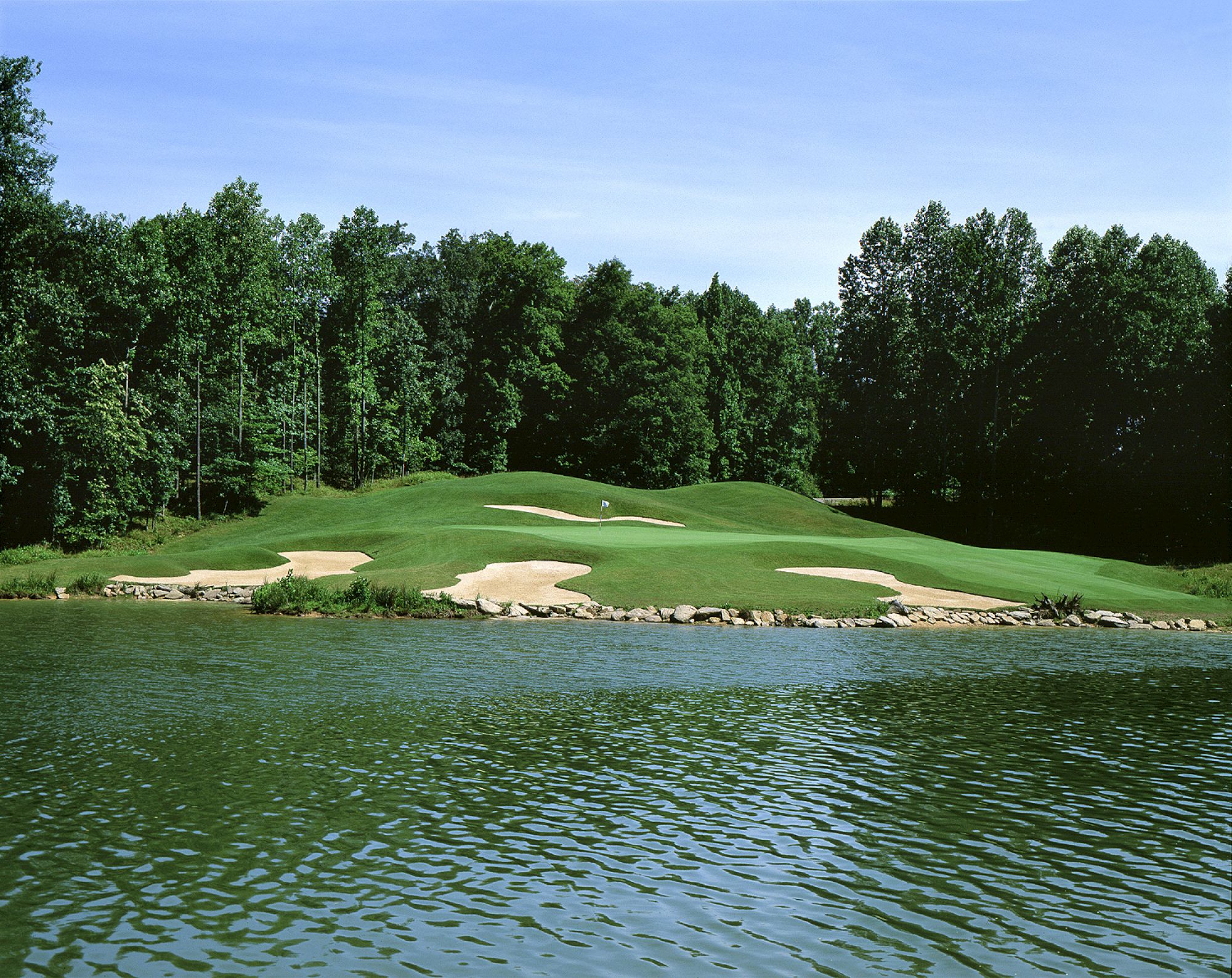 The image shows a golf course with a green area surrounded by trees and a water hazard in the foreground.