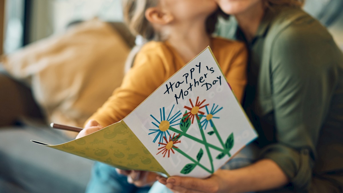 A mother and child share a hug, smiling, holding a Mother's Day card with colorful flowers and 