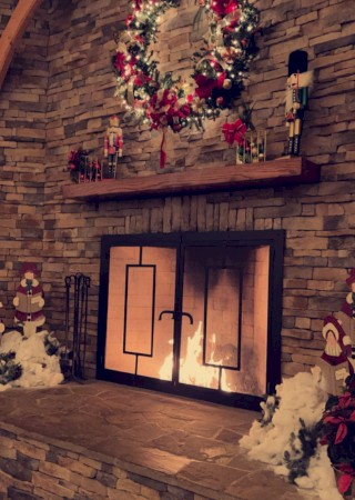 A stone fireplace with glass doors, festive decor: a large wreath above, candles on the mantle, and snowman decor flanking the hearth.