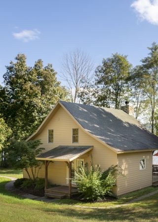 A small house with a covered porch is surrounded by trees and greenery on a sunny day, with a body of water visible in the background.