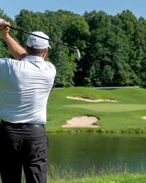 A man in a white shirt and hat swings a golf club on a lush course with water and sand traps, surrounded by dense green trees on a sunny day.