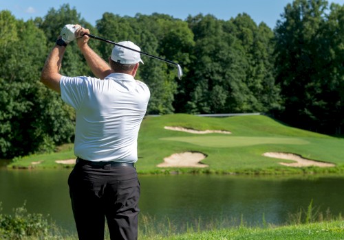 A man in a white shirt and hat swings a golf club on a lush course with water and sand traps, surrounded by dense green trees on a sunny day.