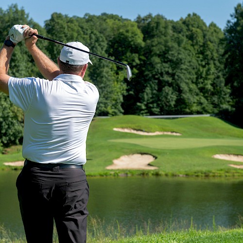 A man in a white shirt and hat swings a golf club on a lush course with water and sand traps, surrounded by dense green trees on a sunny day.