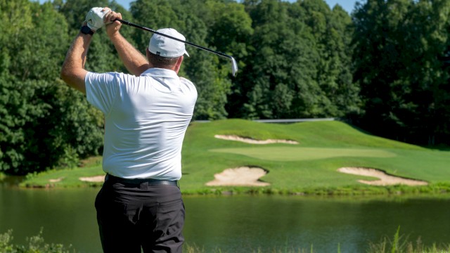 A man in a white shirt and hat swings a golf club on a lush course with water and sand traps, surrounded by dense green trees on a sunny day.