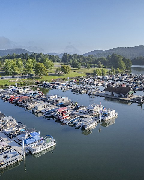 Aerial view of a marina with boats docked on calm water, surrounded by green hills and a parking lot on a clear day.