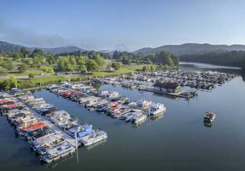 Aerial view of a marina with boats docked on calm water, surrounded by green hills and a parking lot on a clear day.
