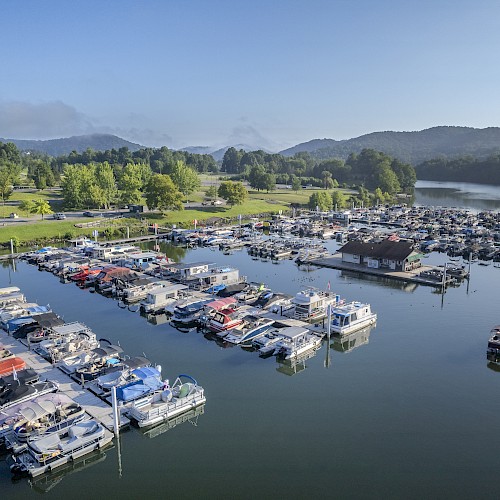 Aerial view of a marina with boats docked on calm water, surrounded by green hills and a parking lot on a clear day.