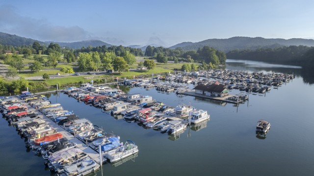 Aerial view of a marina with boats docked on calm water, surrounded by green hills and a parking lot on a clear day.