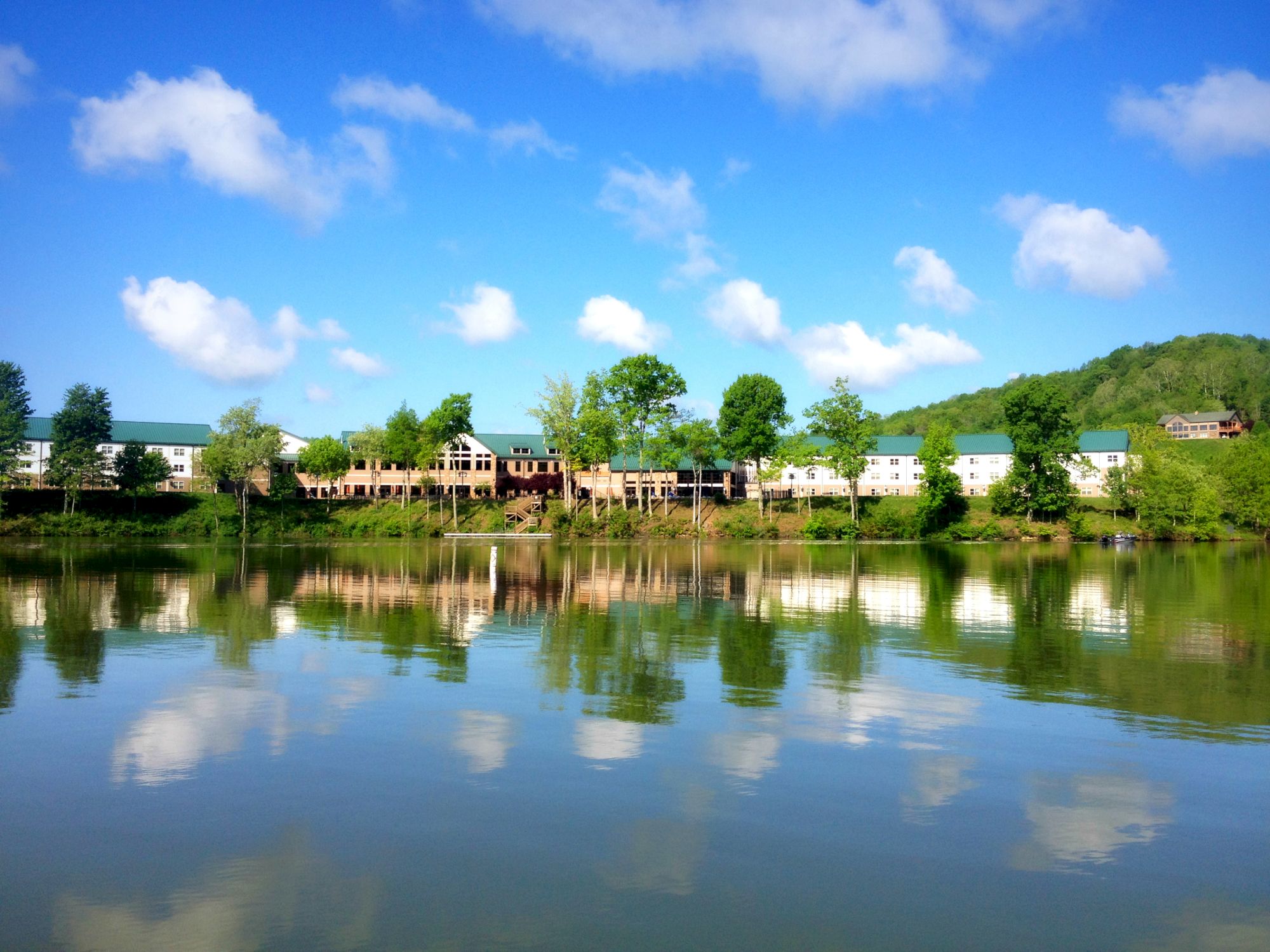 A lakeside view with buildings and trees reflected in the water under a sunny, blue sky with fluffy clouds, ending the sentence.