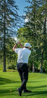 A golfer swings a club on a lush, green golf course under a clear blue sky with trees and hills in the background.