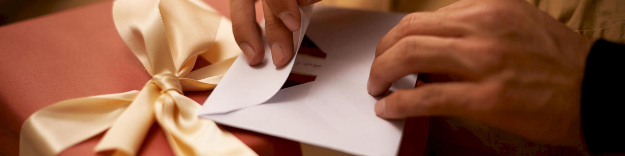 A person is opening an envelope placed on top of a red gift box with a large white ribbon.