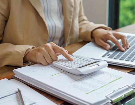 A person is working at a desk with a laptop, calculator, paperwork, and pen, suggesting they are doing financial or business-related tasks.