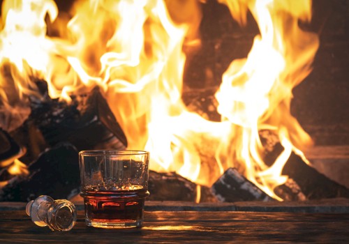 A glass of whisky and a fallen shot glass sit in front of a roaring fire, creating a cozy yet intense scene.
