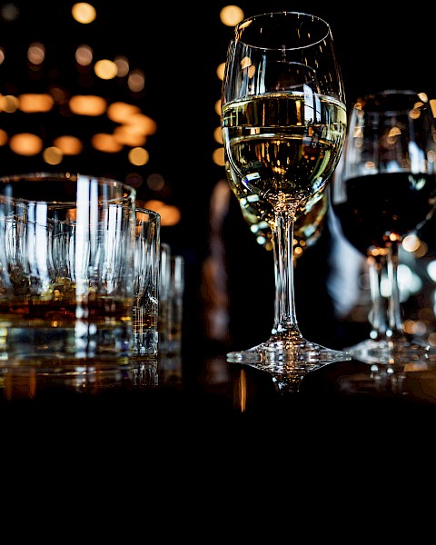 Several glasses of white wine and other beverages are arranged on a dark table with warm bokeh lights in the background.