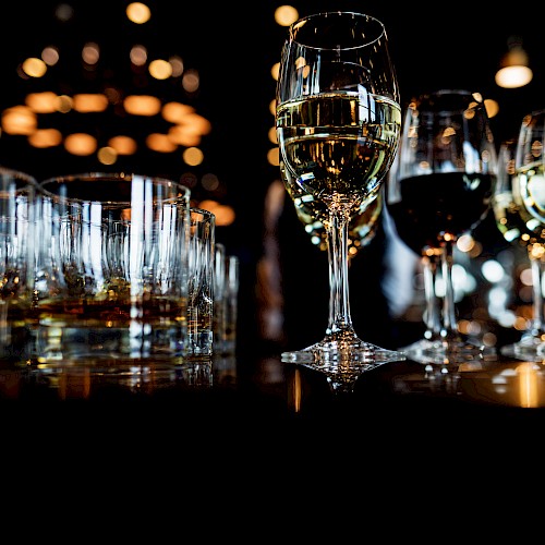 Several glasses of white wine and other beverages are arranged on a dark table with warm bokeh lights in the background.