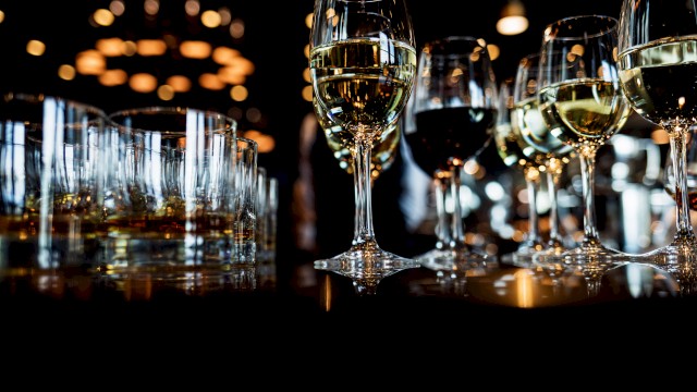 Several glasses of white wine and other beverages are arranged on a dark table with warm bokeh lights in the background.