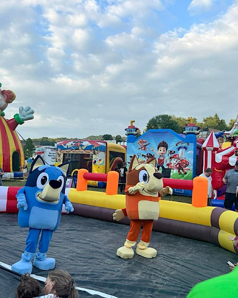 People and children are enjoying a clown-themed inflatable funfair with characters and rides outdoors on a cloudy day.