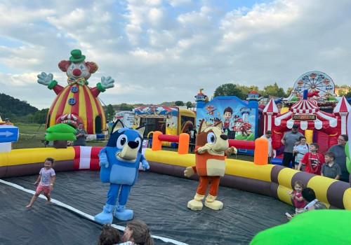 People and children are enjoying a clown-themed inflatable funfair with characters and rides outdoors on a cloudy day.