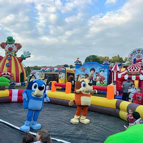 People and children are enjoying a clown-themed inflatable funfair with characters and rides outdoors on a cloudy day.