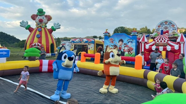 People and children are enjoying a clown-themed inflatable funfair with characters and rides outdoors on a cloudy day.