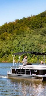 A pontoon boat with two people on a lake, surrounded by green trees and hills, under a clear blue sky during daytime.