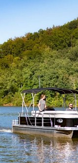 A pontoon boat with two people on a lake, surrounded by green trees and hills, under a clear blue sky during daytime.