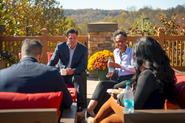 Five people are having a discussion outdoors on a sunny patio with autumn trees in the background.