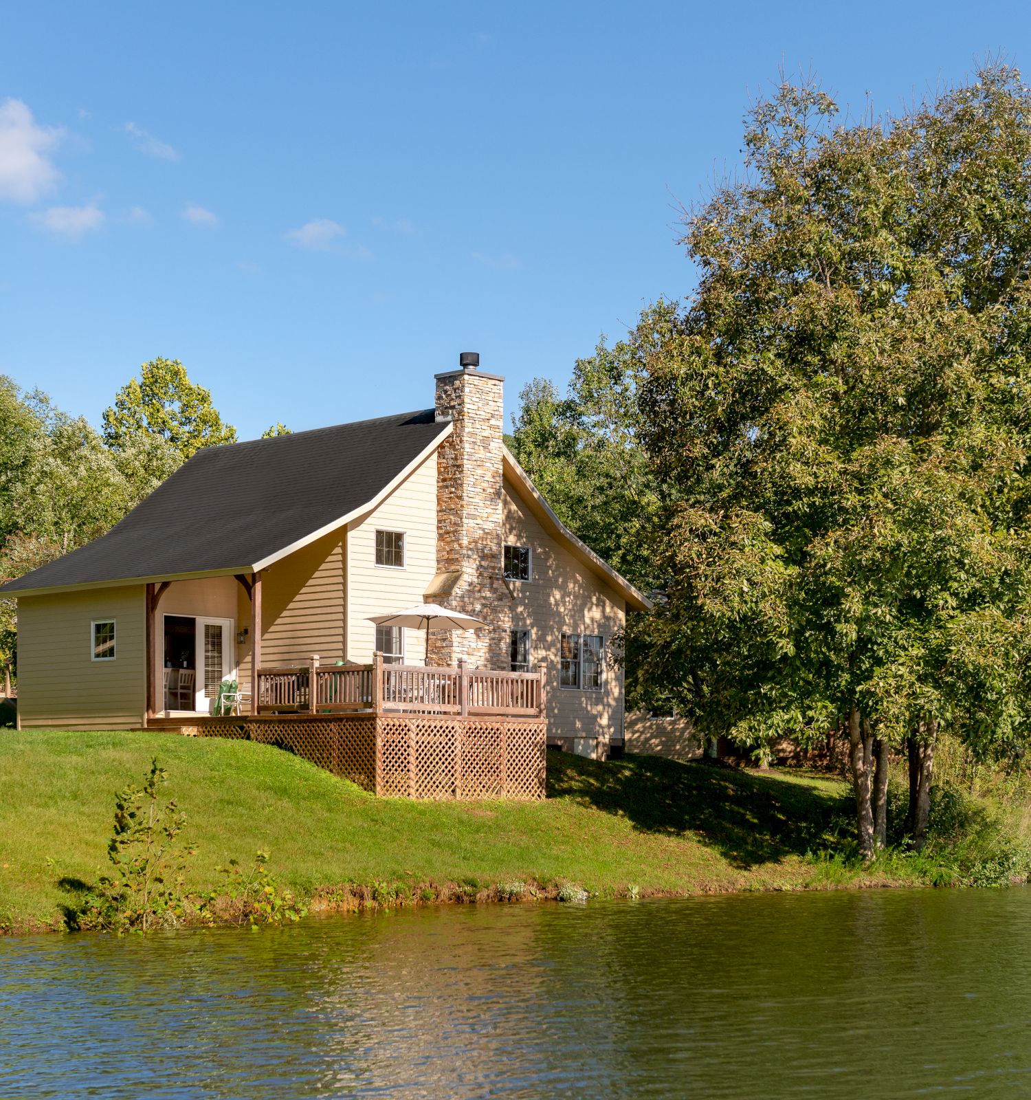 A cozy house with a chimney sits near a lake, surrounded by lush trees under a clear blue sky.