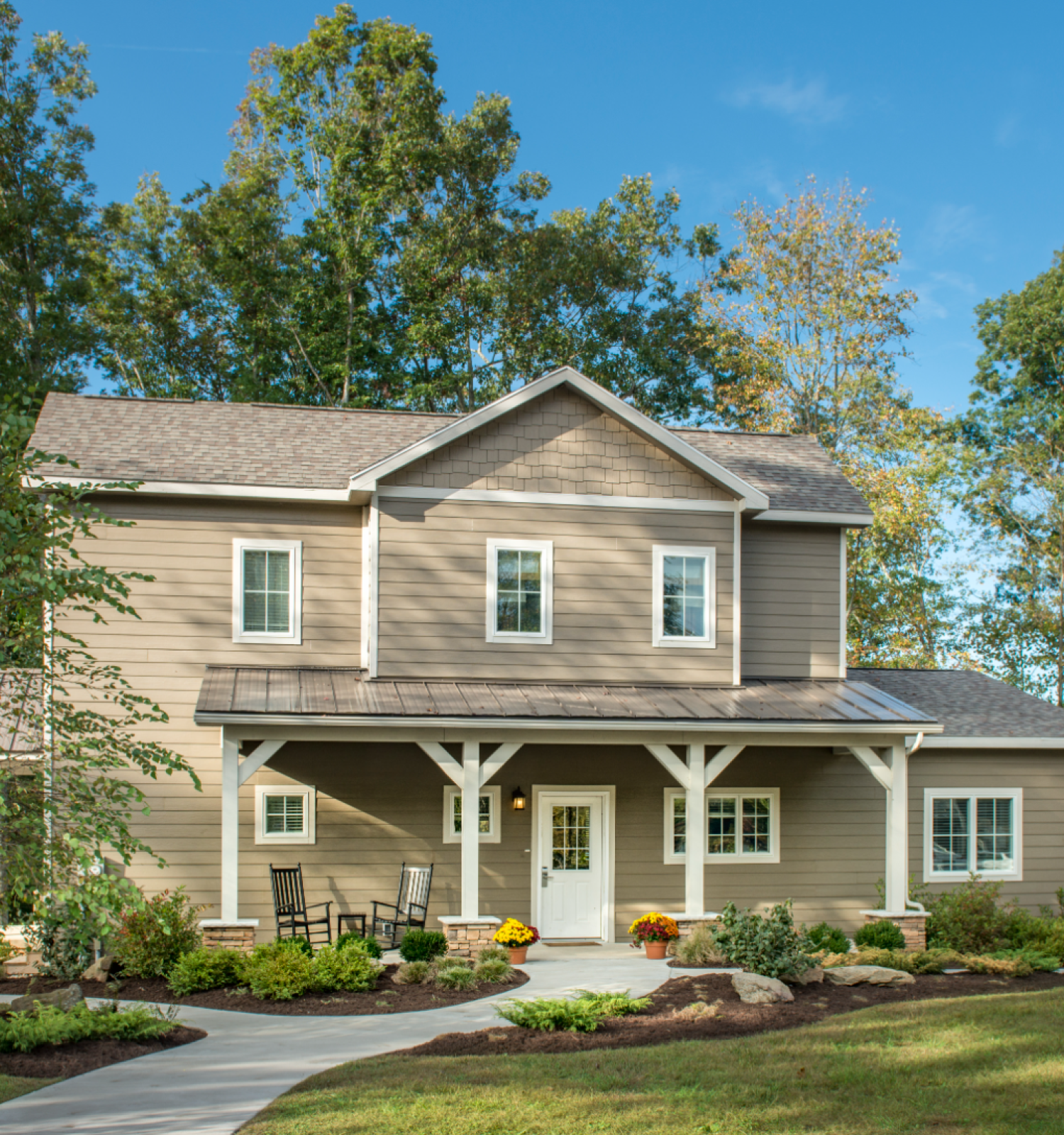 A two-story house with a porch is set in a landscaped yard, surrounded by trees and a clear blue sky.