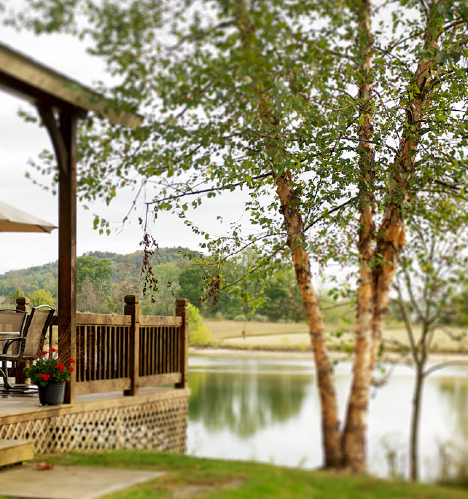 A lakeside porch with a wooden deck, table and chairs under a patio umbrella, lush trees, and calm water in the background, peaceful.