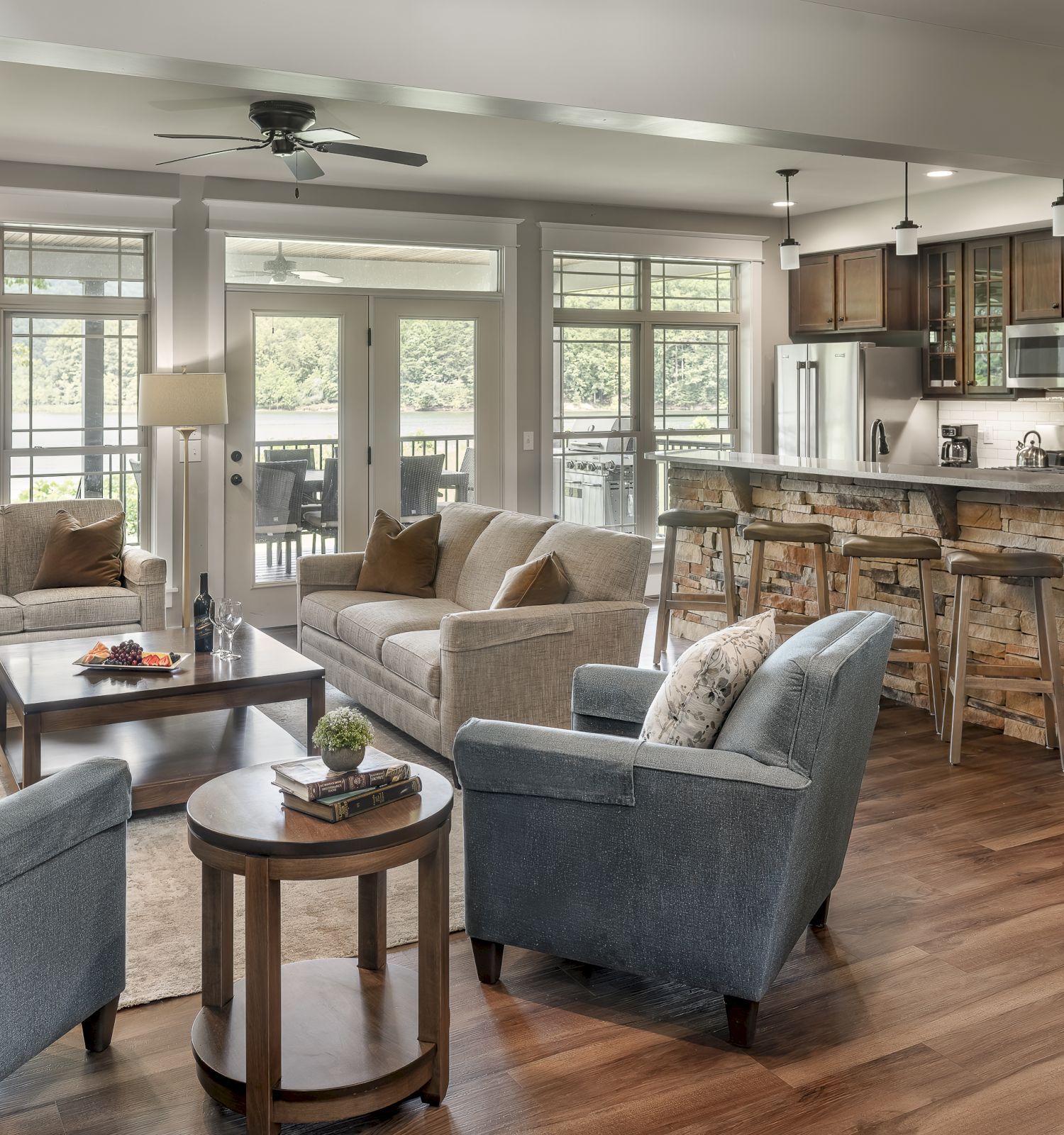 A modern living room and kitchen with hardwood floors, grey and beige furniture, large windows, and bar seating in an open-concept design.