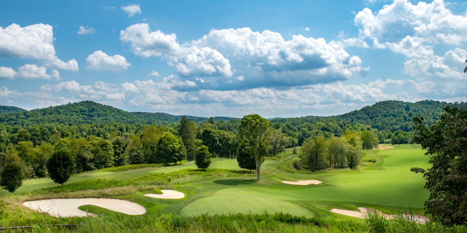 A scenic view of a golf course with lush greenery, sand bunkers, rolling hills, and a partly cloudy blue sky in the background.
