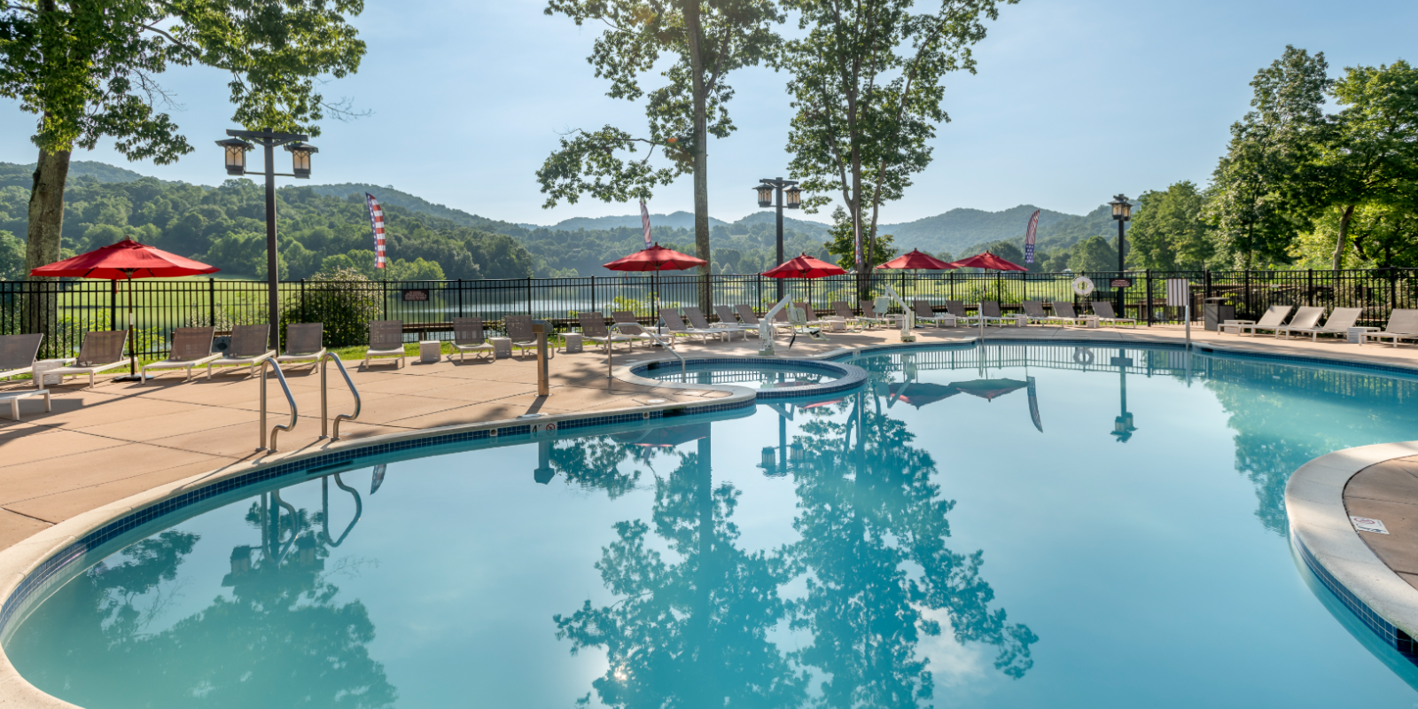 A serene outdoor pool area with clear water, surrounded by trees, lounge chairs, and red umbrellas under a sunny sky.
