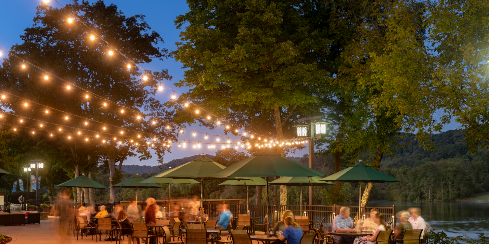 Outdoor dining area by a river with people seated at tables, surrounded by string lights and trees in the evening ambiance.