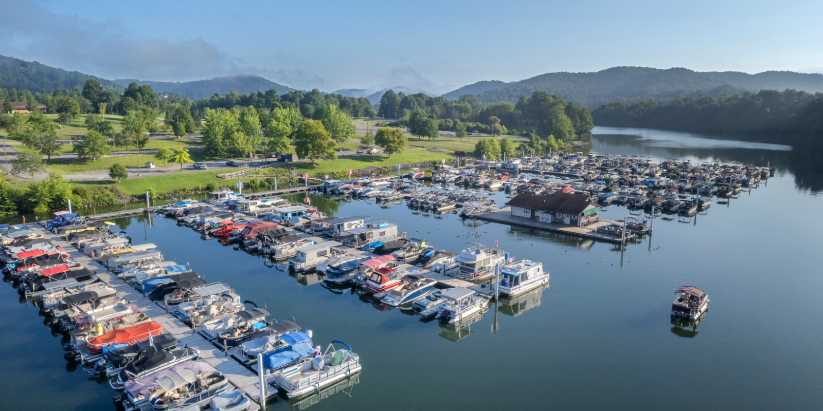 A marina with numerous boats docked on calm water, surrounded by lush green trees and hills under a clear blue sky.
