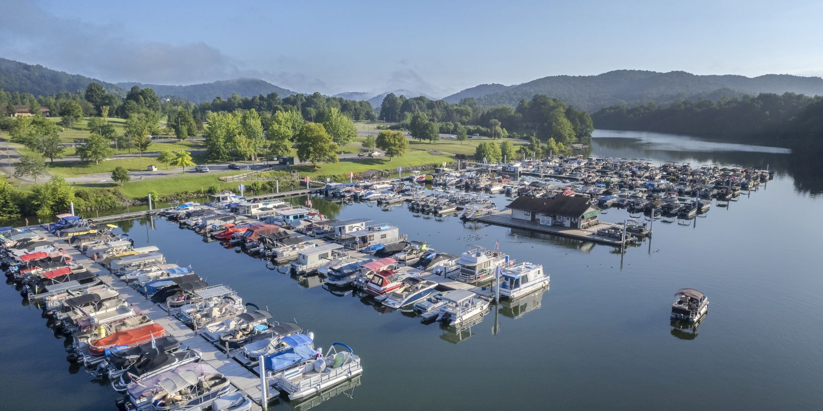 The image shows a marina with numerous boats docked on a calm lake. Surrounding the marina are green hills and a partially wooded area.