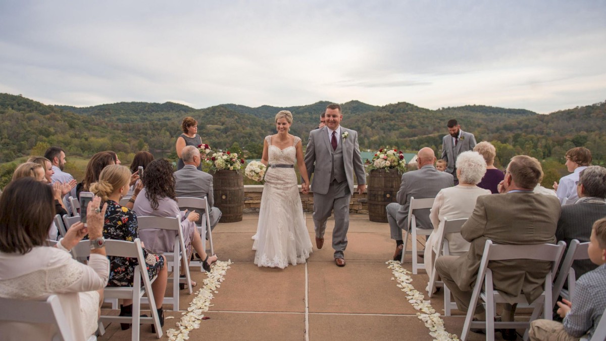 A bride and groom are walking down the aisle outdoors, surrounded by guests sitting on white chairs with a scenic mountain background.