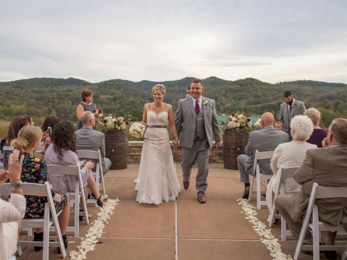 A bride and groom are walking down the aisle outdoors, surrounded by guests sitting on white chairs with a scenic mountain background.