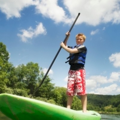 A person is paddleboarding on a green board, wearing a life jacket and red shorts, with a scenic landscape and blue sky in the background.