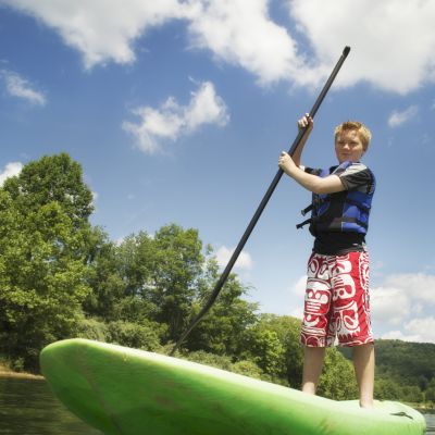 A person is paddleboarding on a calm body of water with trees and hills in the background, wearing a life jacket and red shorts.