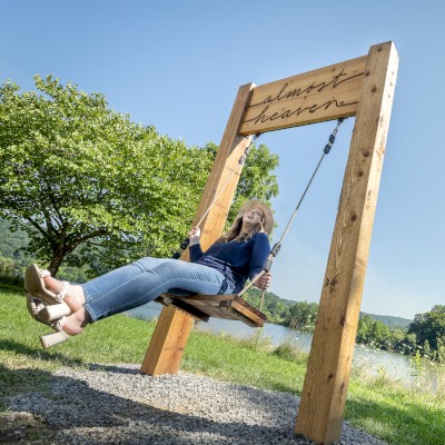 A person is sitting on a wooden swing overlooking a scenic landscape with trees, grass, and a lake under a clear blue sky.