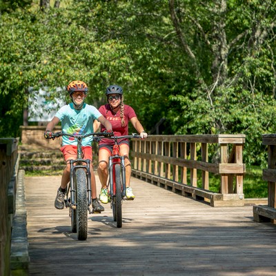 Two people are riding bikes across a wooden bridge surrounded by lush greenery and a sunny atmosphere.