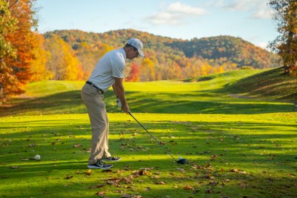 A man is preparing to hit a golf ball on a sunny, scenic golf course surrounded by colorful fall trees.