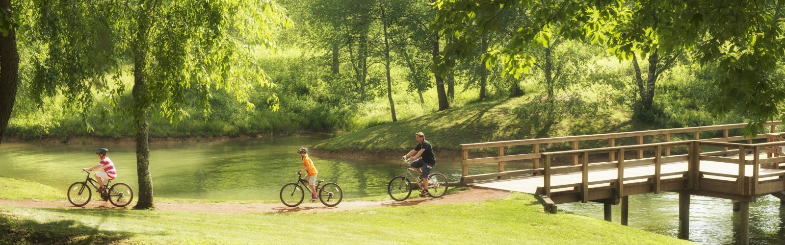 Three people are bicycling on a dirt path through a park with lush greenery and a wooden bridge over water in the background.