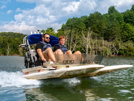 Two people are riding a small, motorized boat on a lake surrounded by lush green trees under a partly cloudy sky.