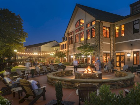 A cozy evening at a lodge with a fire pit, outdoor seating, string lights, and people enjoying the ambiance in front of a large building.