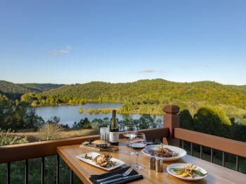 A table set with various dishes, wine, and glasses on a balcony overlooking a serene lake and lush green hills under a clear blue sky, ending the sentence.
